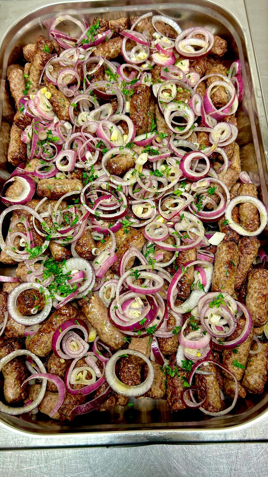 Saftige Cevapcici mit roten Zwiebelringen und frischer Petersilie angerichtet